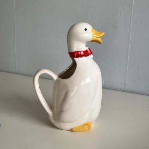 Vit kanna i form av en anka - Charmig vit kanna i form av en anka med gul näbb och fötter samt ett rött band runt halsen. Perfekt för att servera drycker med stil. Tillverkad av keramik och märkt med Falbygdens Ost på undersidan.