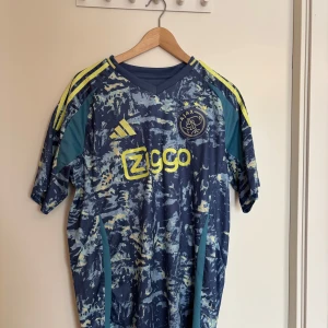 Blå och gul Ajax fotbollströja från Adidas - Säljer en cool Ajax fotbollströja från Adidas i blått med gula detaljer. Tröjan har ett unikt mönster och korta ärmar. Perfekt för fotbollsfans som vill sticka ut på läktaren eller planen. Slim fit design för en modern look.