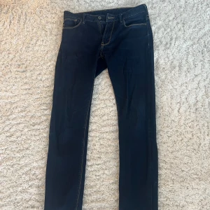 Mörkblå jeans från Emporio Armani - Snygga mörkblå jeans från Emporio Armani med klassisk femficksdesign och knappgylf. Jeansen har en stilren look med kontrastsömmar och en diskret logga på bakfickan. Perfekta för en avslappnad men stilfull outfit. Storlek 30:30