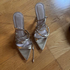 Guldiga klackskor med snörning - Snygga guldiga klackskor med spetsig tå och elegant snörning som går upp runt vristen. Perfekta för en festlig kväll eller speciella tillfällen.