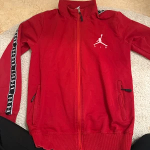 Röd träningsjacka från Jordan - Snygg röd träningsjacka från Jordan med dragkedja och ikonisk logga på bröstet. Jackan har långa ärmar med textdetaljer längs ärmarna och praktiska fickor med dragkedja. Perfekt för sportiga tillfällen eller en casual look.