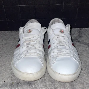 Moncler skor - Snygga vita sneakers från Moncler med klassiska röd och blå ränder på sidan. Skorna har snörning och är tillverkade i skinn, vilket ger en stilren och tidlös look. Perfekta för en avslappnad men ändå elegant stil.