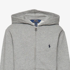 Grå hoodie från Ralph Lauren - Snygg grå hoodie från Ralph Lauren med dragkedja och klassisk logga på bröstet. Perfekt för en avslappnad stil. Den har en huva och ribbade muddar vid ärmslut och nederkant. Helt oanvänd