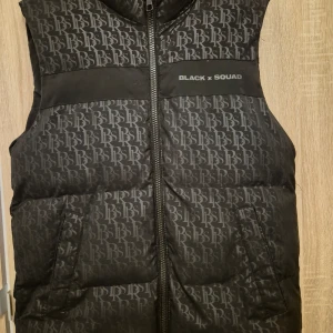 Svart dunväst från Black Squad - Snygg svart dunväst från Black Squad med ett mönster av bokstäverna BSR över hela västen. Västen har en dragkedja framtill och två fickor. Perfekt för en cool och avslappnad stil.