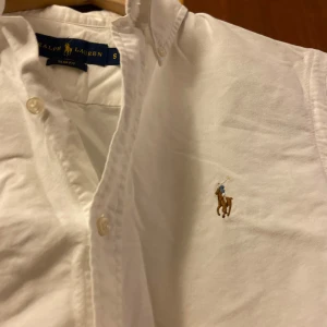 Vit skjorta från Ralph Lauren - Klassisk vit skjorta från Ralph Lauren med broderad logga på bröstet. Skjortan har en slim fit passform och långa ärmar med knappar. Perfekt för en stilren look.