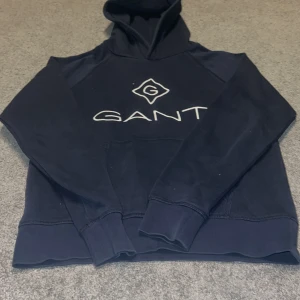 Mörkblå hoodie från GANT - Säljer en mörkblå hoodie från GANT med logga på bröstet. Tröjan har långa ärmar och en klassisk huva. Perfekt för en avslappnad stil. Passar ungdomar i åldern 13-14 år.