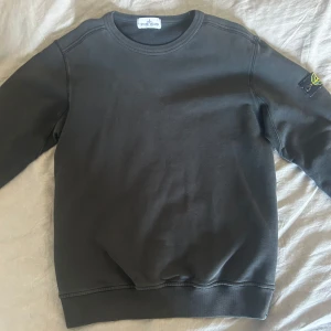 Svart tröja från Stone Island Junior - Säljer en svart tröja från Stone Island Junior. Tröjan har långa ärmar och en rund halsringning. Den är stilren med en logotyp på ärmen.    Storlek 170 (junior)