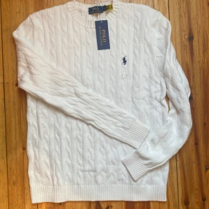 Vit stickad tröja från Polo Ralph Lauren - Säljer en stilren vit stickad tröja från Polo Ralph Lauren med klassiskt flätmönster. Tröjan har en broderad logotyp på bröstet och ribbade muddar vid ärmslut och nederkant. Perfekt för en elegant och avslappnad look.