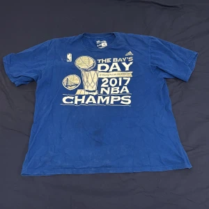 Golden State Warriors 2017 NBA champions tröja - Säljer en blå t-shirt från Adidas med tryck 'The Bay's Day 2017 NBA Champs'. Tillverkad i 100% bomull, perfekt för basketfans. Kortärmad och bekväm passform.