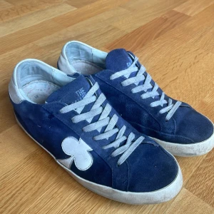 Blå sneakers med vit klöverdetalj - Snygga blå sneakers från The Gallerist med en unik vit klöverdetalj på sidan. Skorna har vita snören och en svart sula. Perfekta för en avslappnad stil. Priset går att diskuteras