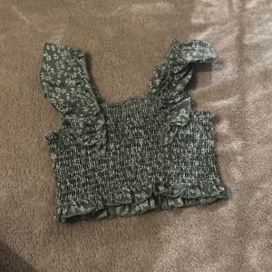 Grön blommig topp från Shein - Säljer en söt grön blommig topp från Shein med smockdetaljer och volanger. Toppen har breda axelband och en croppad passform. Perfekt för varma dagar!