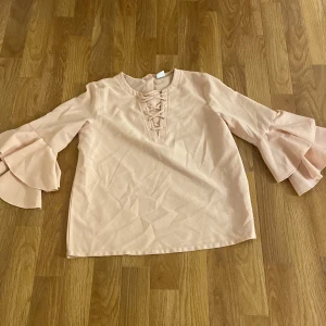 Beige blus från Lindex Kids - Säljer en söt beige blus från Lindex Kids med volangdetaljer på ärmarna och snörning framtill. Perfekt för en stilren och lekfull look. Passar barn i åldern 8-10 år.