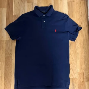 Rosa pikétröja från Polo Ralph Lauren - Snygg rosa pikétröja från Polo Ralph Lauren med klassisk krage och knappar. Den har en broderad blå logga på bröstet. Perfekt för en stilren look.