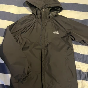 North face jacka (regn/vind) - Säljer en svart regnjacka från The North Face med huva och dragkedja. Jackan har långa ärmar och är perfekt för regniga dagar. Den är stilren och funktionell med logotypen på bröstet.