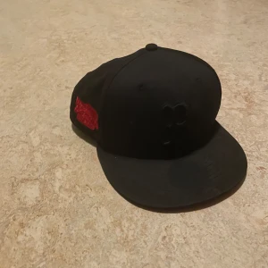Svart/Röd keps från New Era - Snygg svart/Röd keps från New Era med röd broderad detalj på sidan. Kepsen har en platt skärm och är i storlek 7 3/4 (61,5 cm). 
