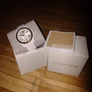 Vit klocka från Michael Kors - Elegant vit klocka från Michael Kors med romerska siffror på urtavlan. Klockan har en stilren design med en vit rem och guldfärgade detaljer. Perfekt accessoar för att lyfta din stil.