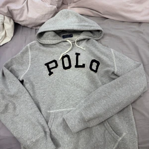 Grå hoodie från Polo Ralph Lauren - Säljer en stilren grå hoodie från Polo Ralph Lauren med svart text på bröstet. Den har en klassisk design med huva och dragsko samt en stor ficka framtill. Perfekt för en avslappnad look.
