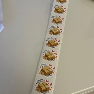 Klistermärken med texten 'Handle with Care' 10st - Säljer en rulle med klistermärken som har texten 'Handle with Care' och 'Fragile'. Klistermärkena har en bild av en kartong och små röda hjärtan runtom. Perfekt för att märka paket som behöver extra varsam hantering. 10 st