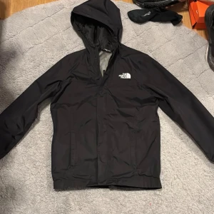 Svart vindjacka från The North Face - Säljer en svart vindjacka från The North Face med huva och dragkedja. Jackan har en enkel design med logotyp på bröstet och är perfekt för blåsiga dagar. Sitter som en S 
