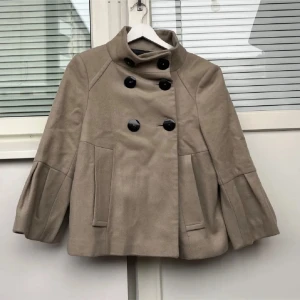 Beige kappa med knappar Zara - Snygg beige kappa med stora svarta knappar och hög krage. Jackan har en unik design med veckade detaljer på ärmarna och en dubbelknäppning framtill. Perfekt för att ge en stilren look.