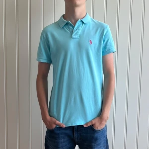 Ljusblå pikétröja från Polo Ralph Lauren - Säljer en stilren ljusblå pikétröja från Polo Ralph Lauren. Tröjan har en klassisk krage och korta ärmar, med den ikoniska loggan broderad i rosa på bröstet. Perfekt för en avslappnad och snygg look. Storleken är S och skicket är mycket bra.