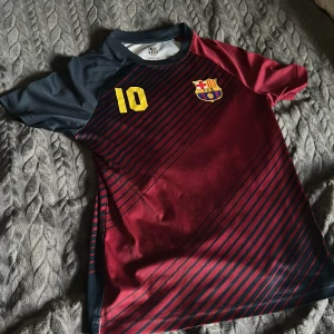 FC Barcelona fotbollströja - Snygg FC Barcelona fotbollströja i röd och mörkblå med gula detaljer. Tröjan har korta ärmar och ett randigt mönster. Nummer 10 och klubbens emblem pryder framsidan, medan baksidan har texten 'FC BARCELONA'. Perfekt för fans! 💙❤️