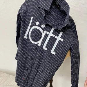 Svart och vit randig skjorta med text - Snygg svart skjorta med vita ränder och stor text 'lätt' på framsidan. Skjortan har långa ärmar och knappar framtill. Perfekt för en avslappnad stil med en twist.