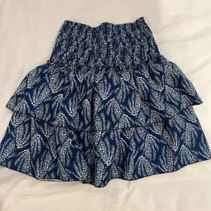 Blå och vit mönstrad kjol - Snygg blå och vit kjol med ett unikt mönster. Kjolen har en volangdetalj och en elastisk midja för en bekväm passform. Perfekt för att ge din outfit en färgklick!