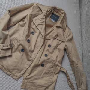 Säljer en snygg beige trenchcoat med knappar och bälte i midjan. Jackan har en klassisk design med långa ärmar och en stilren krage. Perfekt för höst och vår! Köpt second hand men känns helt ny coh oanvänd, ända är att en knapp har trillat av(bild 2)