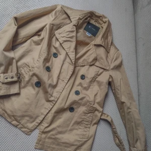 Beige trenchcoat med bälte - Säljer en snygg beige trenchcoat med knappar och bälte i midjan. Jackan har en klassisk design med långa ärmar och en stilren krage. Perfekt för höst och vår! Köpt second hand men känns helt ny coh oanvänd, ända är att en knapp har trillat av(bild 2)