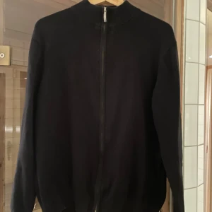 Svart kofta från GANT - Säljer en stilren svart kofta från GANT i storlek L. Den har en dragkedja framtill och är perfekt för både höst och vinter. Koftan är långärmad och har en ribbad krage 