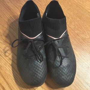 Svarta fotbollsskor med rosa detaljer - Säljer ett par svarta fotbollsskor från Puma i storlek dam 35. Skorna har en tight passform med snörning och en stickad del runt ankeln för extra stöd. De har snygga rosa detaljer som ger en cool kontrast. Perfekta för fotbollsträning eller match!