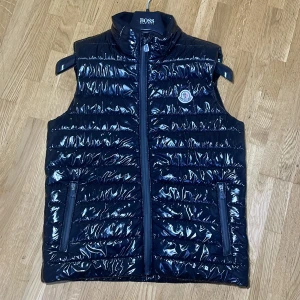 Svart glansig dunväst från Moncler - Säljer en snygg svart glansig dunväst från Moncler. Västen har en dragkedja framtill och två praktiska fickor med dragkedjor. Den är perfekt för höst och vår, och ger en stilren look. Moncler-loggan syns tydligt på bröstet. Passar både till vardags och lite finare tillfällen.