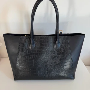 Svart axelväska med krokodilmönster - Säljer en stilren svart axelväska med krokodilmönster. Väskan är rymlig och perfekt för både vardag och jobb. Den har två handtag och är gjord i ett snyggt skinnmaterial. Passar till alla säsonger och ger en elegant touch till din outfit.