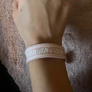 Rosa armband från Christian Dior - Säljer ett jättefint rosa armband som är vävt med texten 'Christian Dior' och har en justerbar knytning med tofsar. Köpte den i somras på en marknad i Spanien så förmodligen är den inte äkta, därför priset. Ett äkta kostar runt 4000