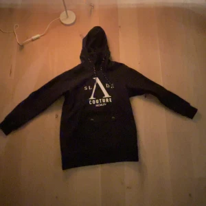 Svart hoodie från Slade Couture - Säljer en svart jack-hoodie från Slade Couture med tryck framtill. Den har en klassisk design med justerbar huva och långa ärmar. Perfekt för en avslappnad stil i backen 