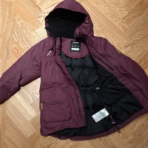 Vinröd vattentät dunparkas Serkkula från Reima  - Säljer en vinröd vinterjacka från Reima med huva och dragkedja. Jackan har en svart insida och är perfekt för kalla dagar. Den är designad för att hålla värmen och har praktiska detaljer som justerbar midja.