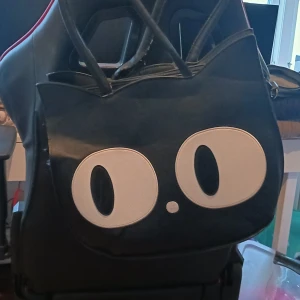 Svart axelväska med kattmotiv - Cool svart axelväska med ett unikt kattmotiv på framsidan. Väskan har två handtag och en justerbar axelrem för bekväm användning. Perfekt för att ge en lekfull touch till din outfit.