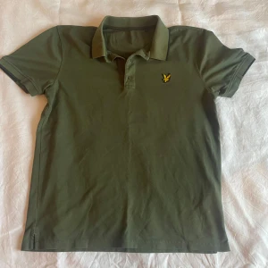 Grön pikétröja från Lyle & Scott - Snygg grön pikétröja från Lyle & Scott med klassisk krage och knappslå. Tröjan har en broderad logotyp i gult på bröstet. Perfekt för en avslappnad stil.