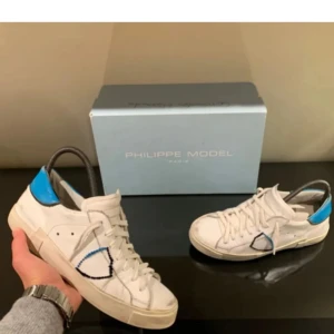 Vita sneakers från Philippe Model  - Snygga vita sneakers från Philippe Model med blå detaljer och snörning. Perfekta för en stilren look. Kan gå ner i pris vid snabb affär, bara att höra av er vid frågor. Inga större hål eller slitningar,Box förekommer 