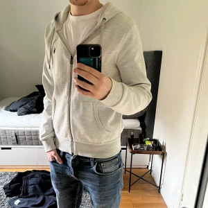 Grå hoodie med dragkedja - Säljer en stilren grå hoodie med dragkedja och huva. Tröjan har långa ärmar och en liten broderad logga på bröstet. Perfekt för en avslappnad look. Raffe hoodie perfekt skick. DM för frågor. Äkta.
