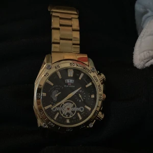 Guldfärgad klocka med svart urtavla - Snygg guldfärgad klocka med svart urtavla och flera små urtavlor. Klockan har en robust länkarmband och detaljer som tachymeter på ringen. Perfekt för den som gillar en elegant och iögonfallande stil.