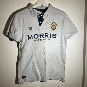 Grå pikétröja från Morris R. Club - Snygg grå pikétröja från Morris R. Club med broderade detaljer och texten 'Morris Rowing Club' på framsidan. Tröjan har en klassisk krage med knappar och korta ärmar. Perfekt för en avslappnad stil.