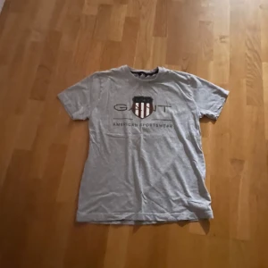 Grå t-shirt från Gant - Säljer en grå t-shirt från Gant med tryck på framsidan. Den har en klassisk passform och korta ärmar, perfekt för en avslappnad stil.