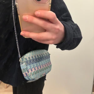 Mönstrad axelväska med dragkedja Becksöndergaard - Snygg axelväska med ett unikt vågigt mönster i blått, rosa och grönt. Väskan har en praktisk dragkedja och en kedja som axelrem. Perfekt för att ge din outfit en färgglad touch! Från Becksöndergaard i nyskick!