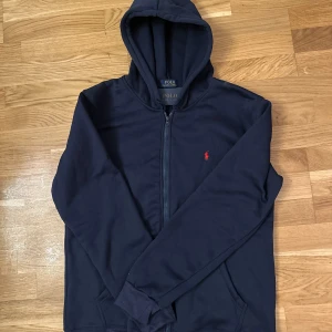 Mörkblå hoodie från Polo Ralph Lauren - Säljer en stilren mörkblå hoodie från Polo Ralph Lauren med dragkedja och den ikoniska röda loggan på bröstet. Perfekt för en avslappnad look.