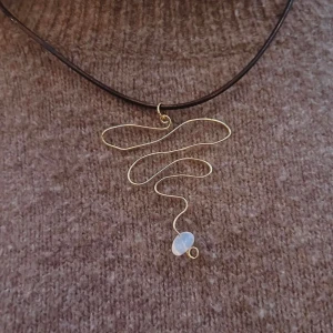 Halsband med guldtråd och opalit - Unikt halsband med en slingrande design i guldtråd och en vit pärla. Hängs på en tunn brun rem i äkta läder. Perfekt för att ge en elegant touch till din outfit.