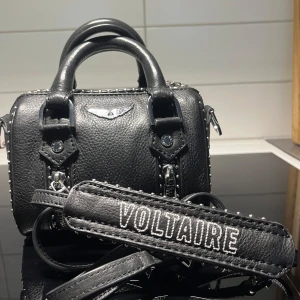 Svart handväska från Voltaire - Snygg svart handväska i skinn med coola detaljer som kedjor och nitar. Väskan har ett handtag och en avtagbar axelrem med texten 'VOLTAIRE'. Perfekt för att ge din outfit sista touchen. Det är en Zadig väska, i minsta storleken. Den är äkta. Jag köpte den ny för 3999kr på NK.