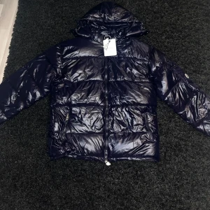 Marinblå pufferjacka från Moncler - Snygg svart pufferjacka från Moncler med glansig finish och huva. Jackan har en dragkedja framtill och en praktisk ficka med Moncler-logga på ärmen. Perfekt för kyliga dagar.                   PRIS KAN DISKUTERAS!