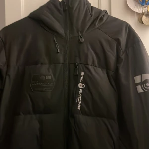 Svart dunjacka från Sail Racing - Snygg svart dunjacka från Sail Racing med huva och dragkedja. Jackan har en logotyp på bröstet och ärmen, samt en praktisk bröstficka med dragkedja. Perfekt för kalla dagar.